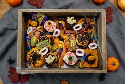 Halloween treat box filled with cupcakes decorated with ghosts, pumpkins, spider webs, and assorted candies in a rustic wooden crate, surrounded by autumn leaves and pumpkins