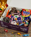 Festive Late Late Toy Show Treat Box filled with Tayto crisps, Dairy Milk bars, candy canes, iced biscuits, and Christmas treats from Hannah’s Kitchen Cork, surrounded by gifts, fairy lights, and a cup of hot chocolate with marshmallows.